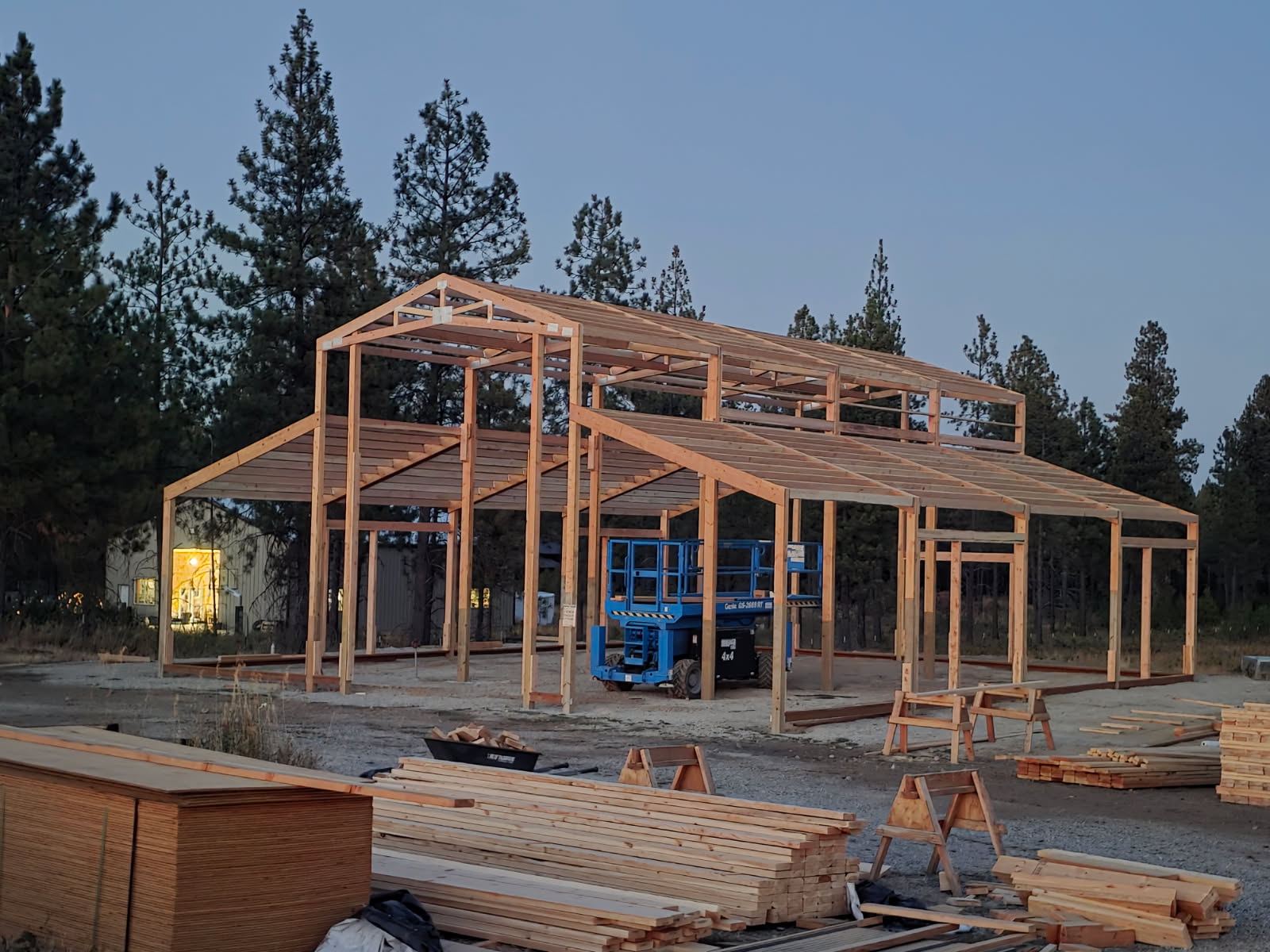 Timber post frame of a barn standing at dusk, with lumber and plywood stacked on site